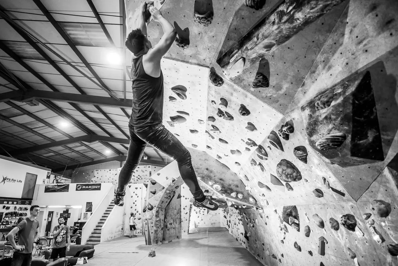 Person climbing an indoor rock wall with various handholds, observed by others below in an indoor climbing facility.