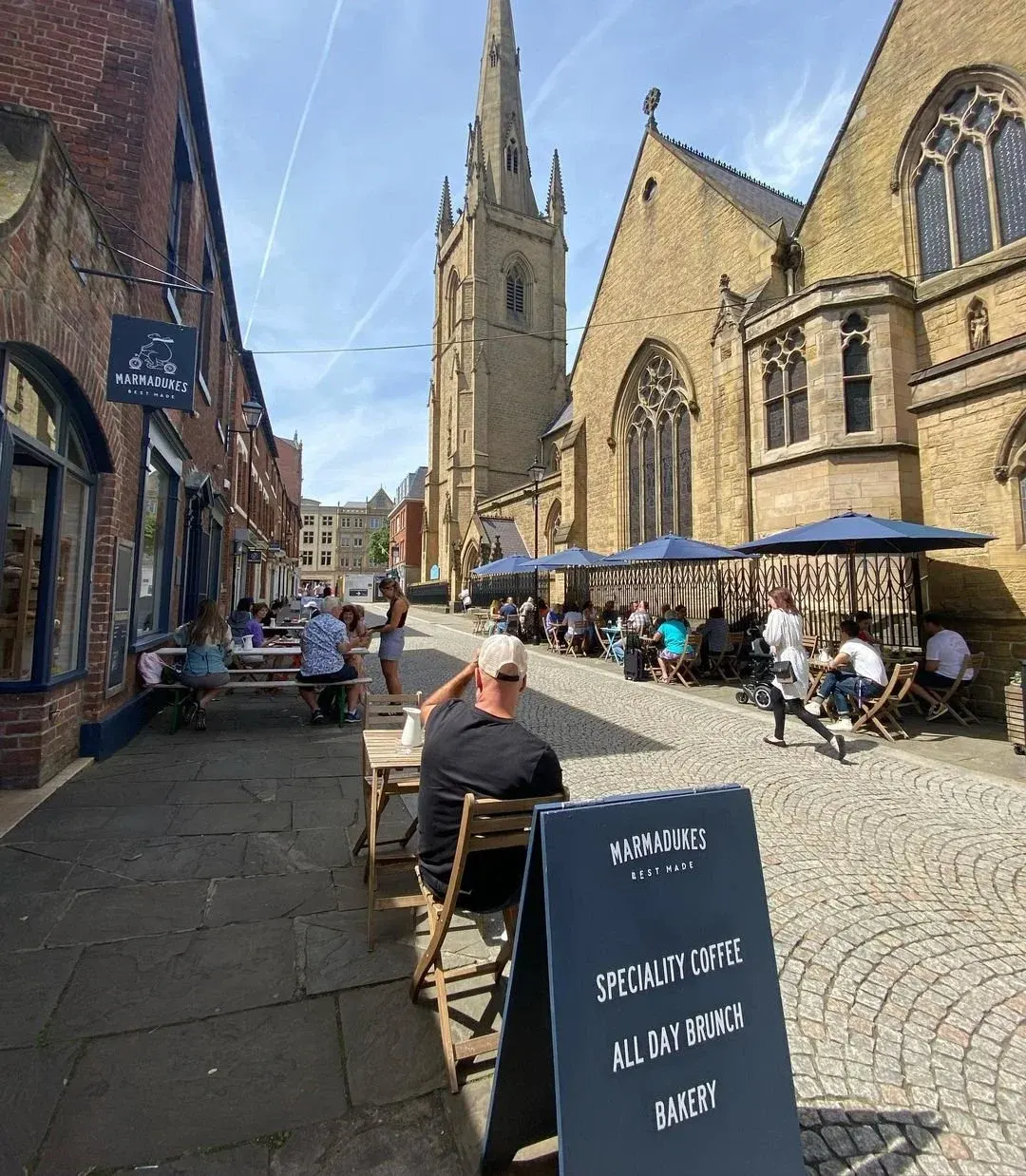 Outdoor café seating on a pedestrianised street lined with historic brick and stone buildings, with a church spire in the background and customers dining under blue umbrellas.