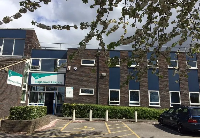 Exterior of a brick library building with teal signage, car park and climbing ivy overhead.