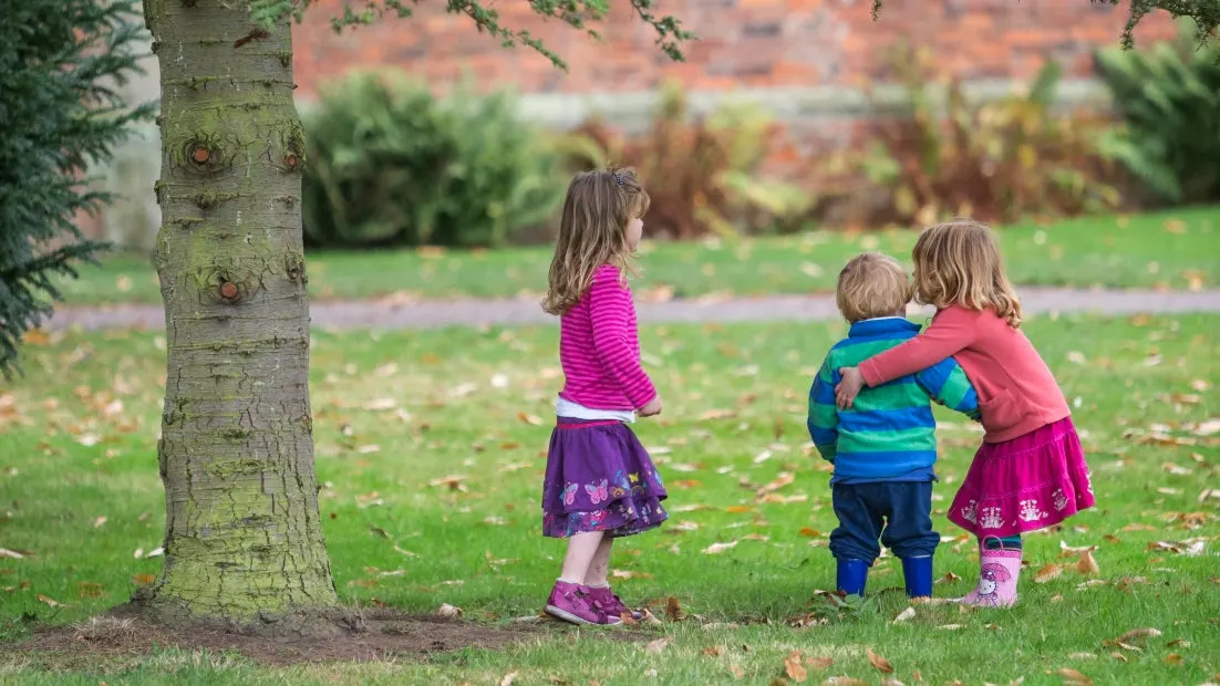 Three young children playing together on a grass lawn beside a large tree, with autumn foliage visible in the background.