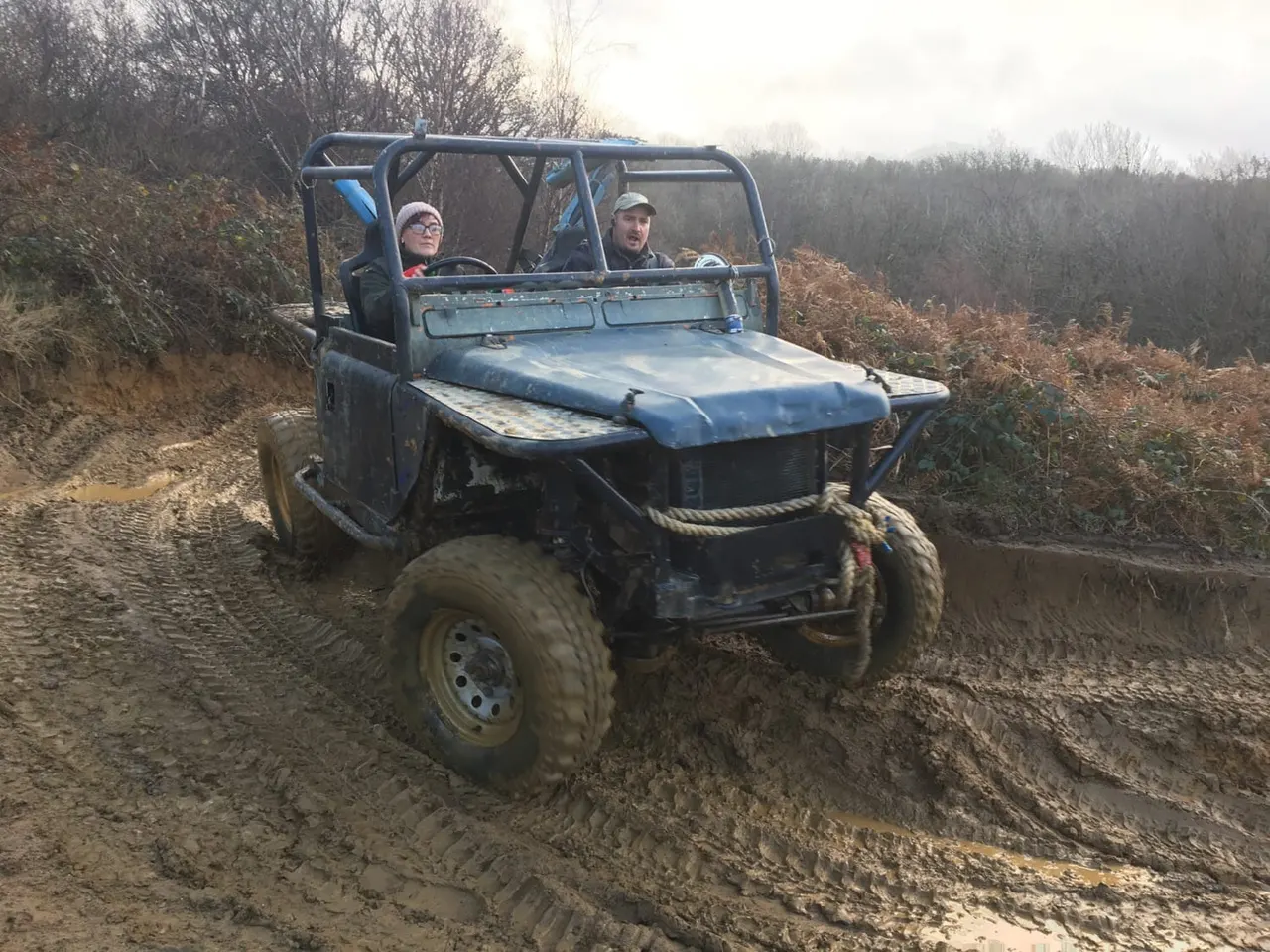 Two people driving a blue off-road buggy through muddy terrain surrounded by bare trees.
