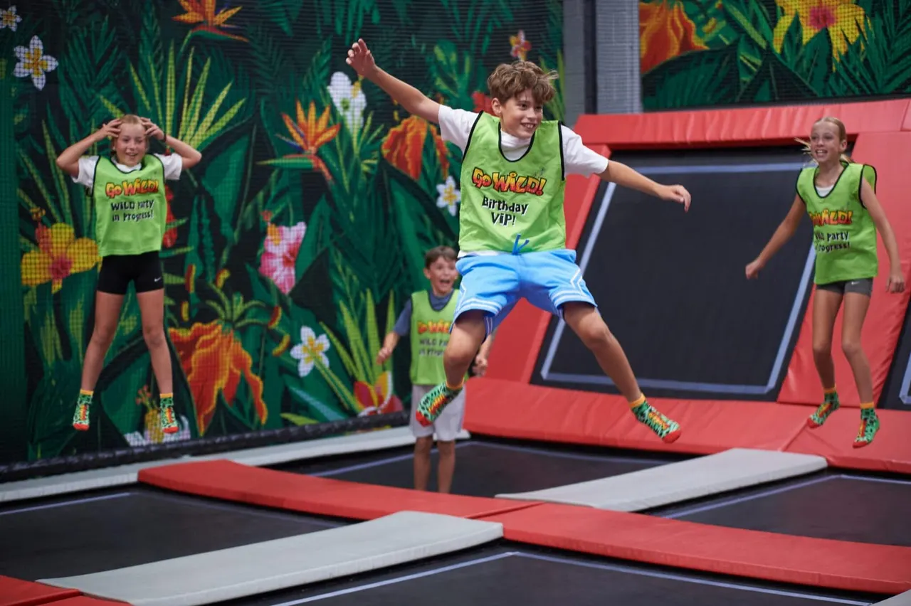 Children in matching neon vests bouncing on a trampoline with a tropical-themed backdrop.