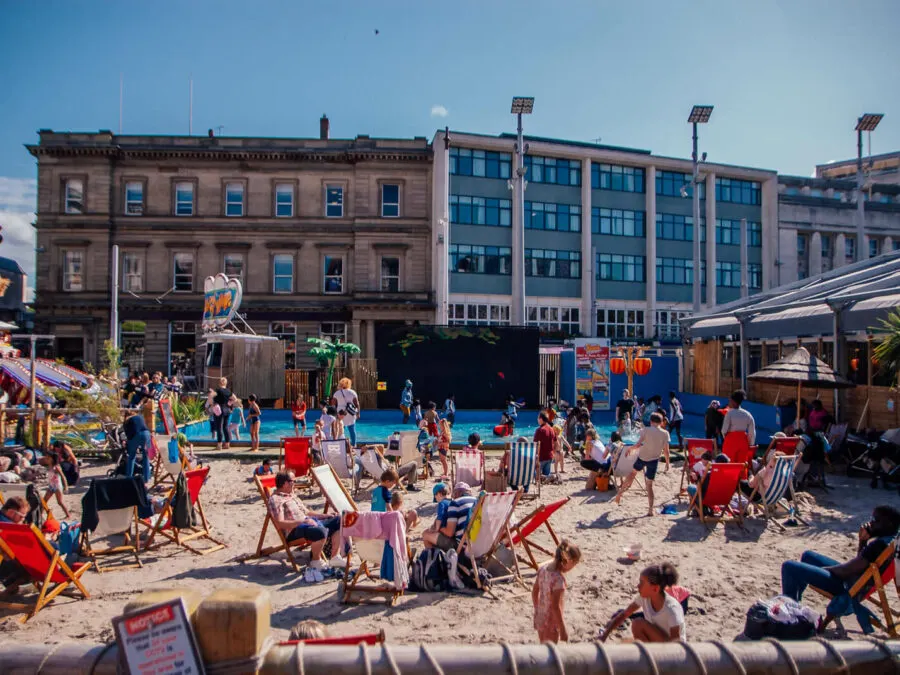 Busy urban beach with visitors relaxing on sand, paddling pool, and Victorian buildings in background under clear blue sky.