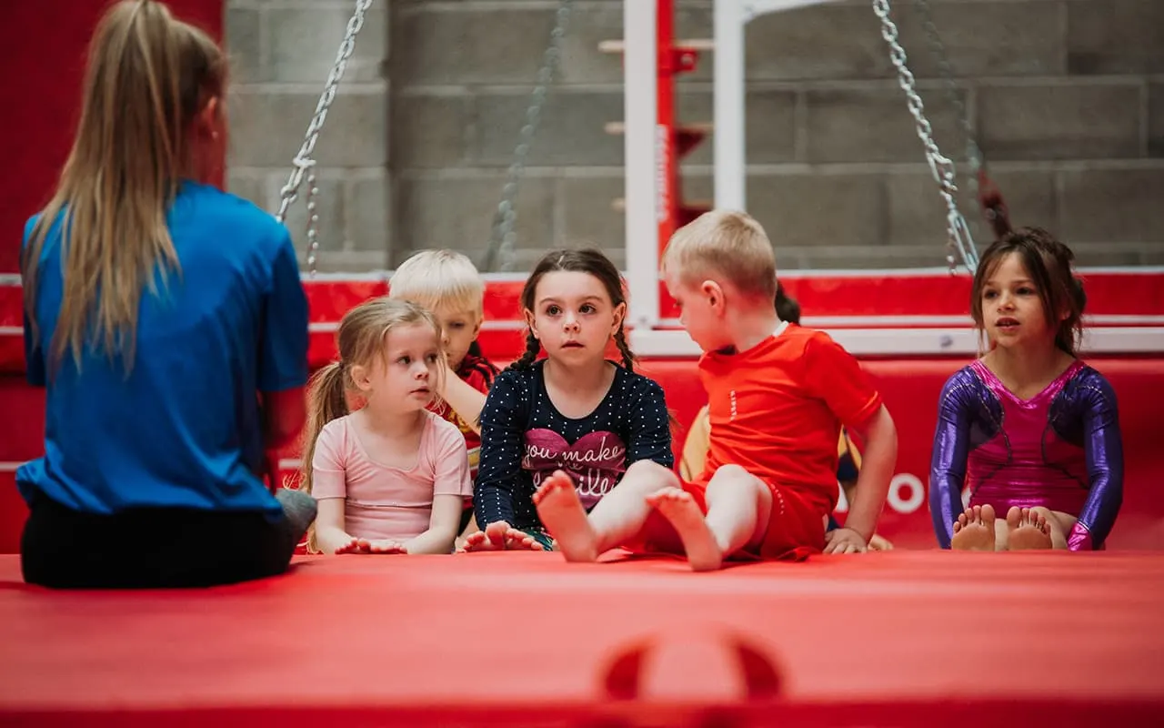 Young children sitting on a red padded floor with an instructor during a gymnastics or movement class.