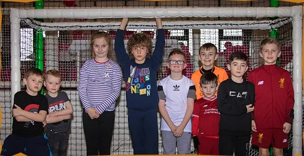 A group of children of various ages standing together in front of a large net goal, smiling at the camera.