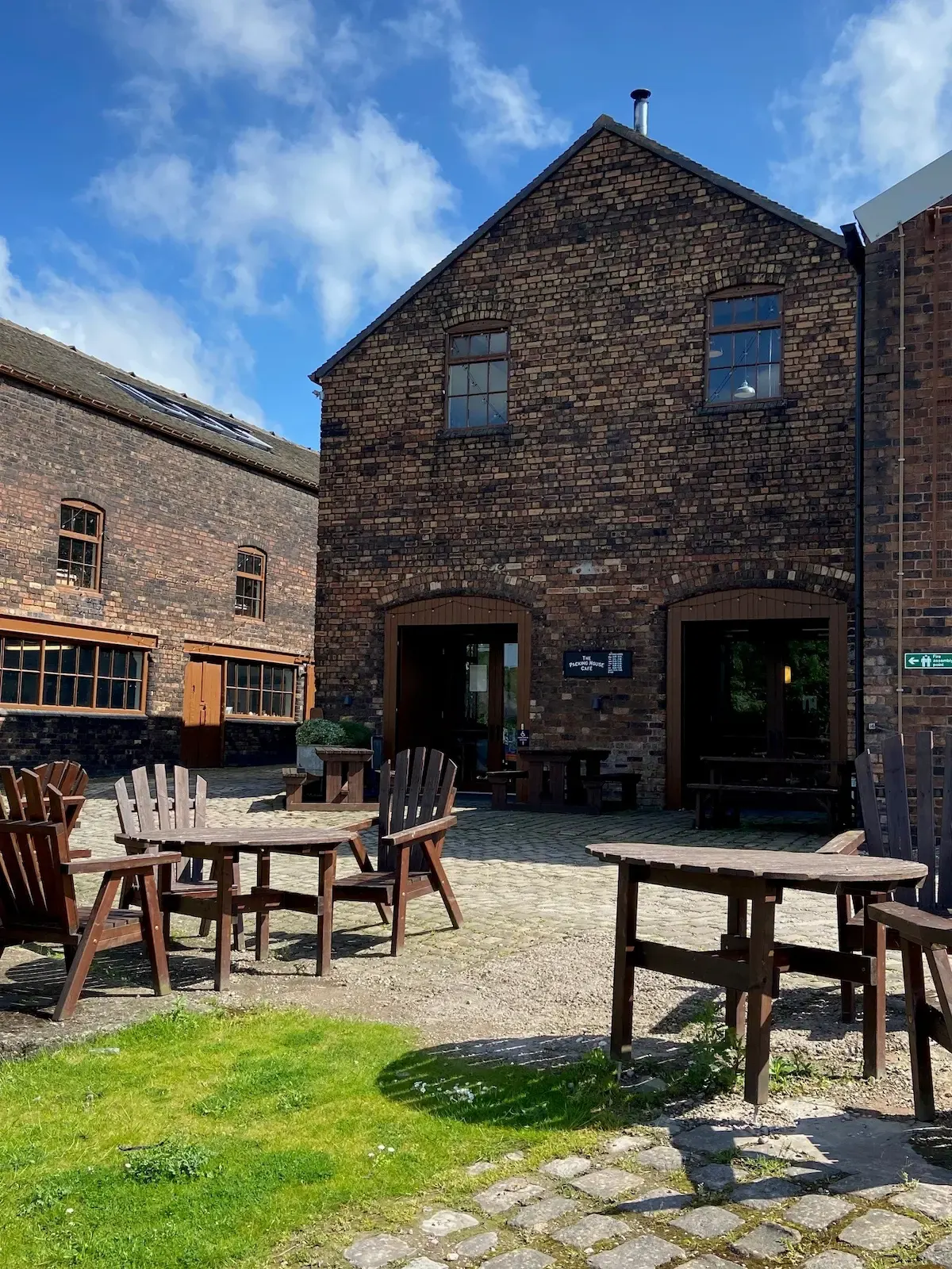 Outdoor courtyard with wooden tables and Adirondack chairs in front of converted brick industrial buildings under blue sky.