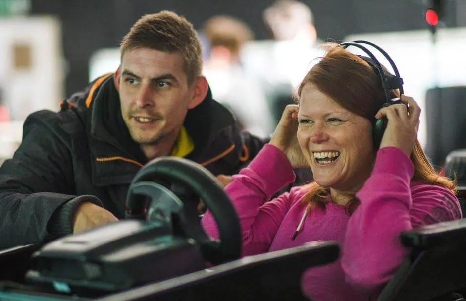 A man and woman wearing headsets smile whilst sitting in an indoor go-kart racing vehicle.