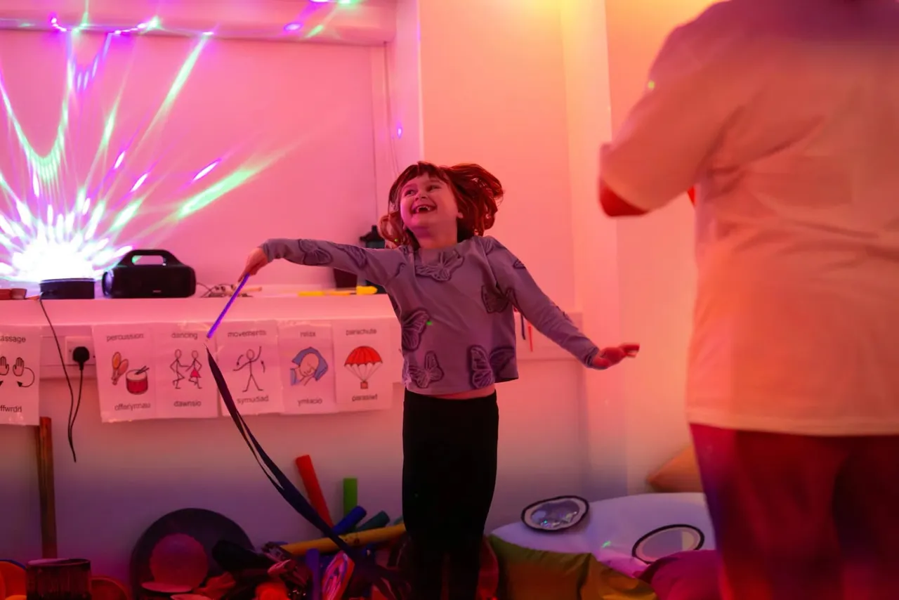 Young child playing in an interactive sensory room with neon pink and orange lighting, surrounded by musical instruments and educational visual displays.