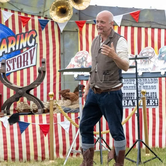 Man in period costume gestures whilst speaking at a circus-themed outdoor performance stage with striped backdrop and vintage signage.