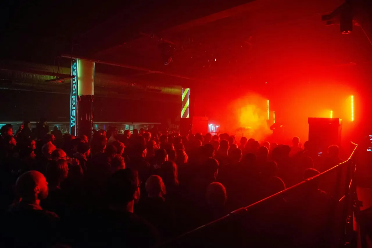 Large crowd dancing on a dancefloor illuminated by red and yellow stage lighting at a nightclub.