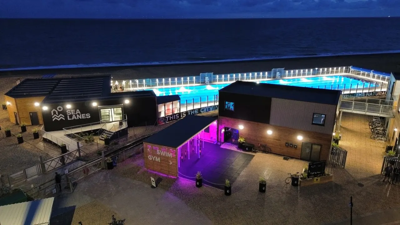 Aerial night view of a waterfront leisure complex with an illuminated swimming pool, gym building with magenta lighting, and Sea Lanes building beside calm water.