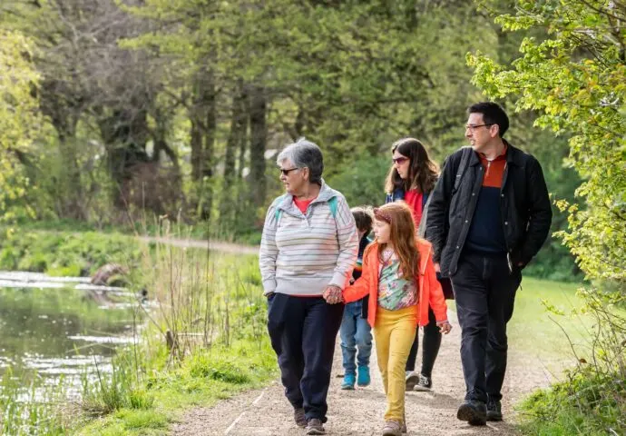 Family of five walking along a tree-lined path beside a still water body in woodland.