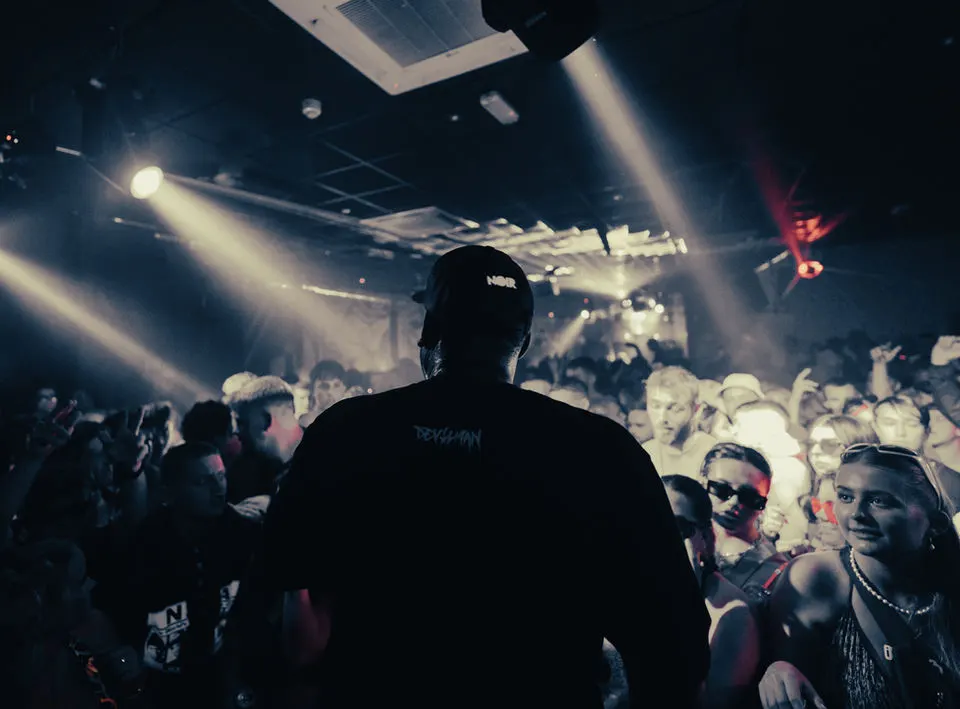 DJ performing at a nightclub with dramatic stage lighting and a crowd of people dancing.