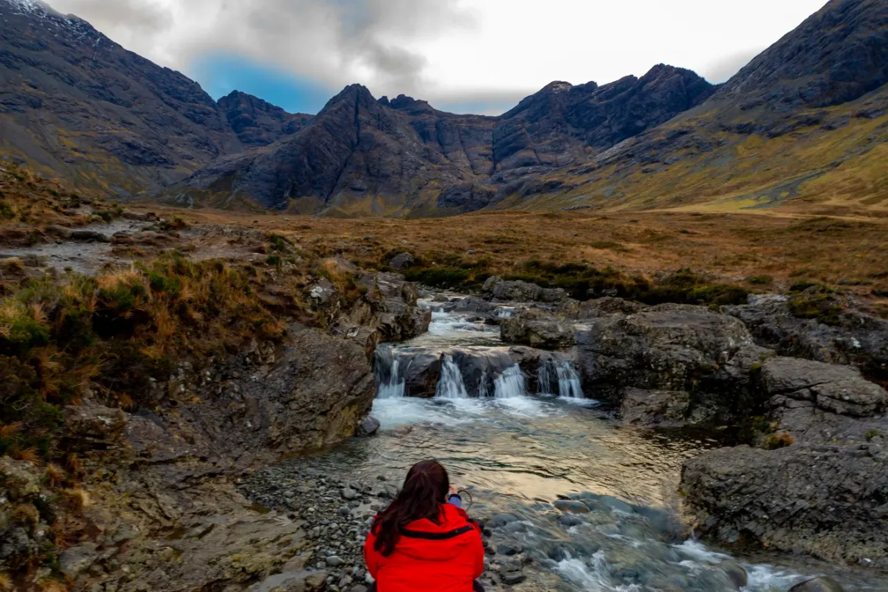 Person in red jacket viewing mountain stream with small waterfalls, surrounded by moorland and rocky peaks.