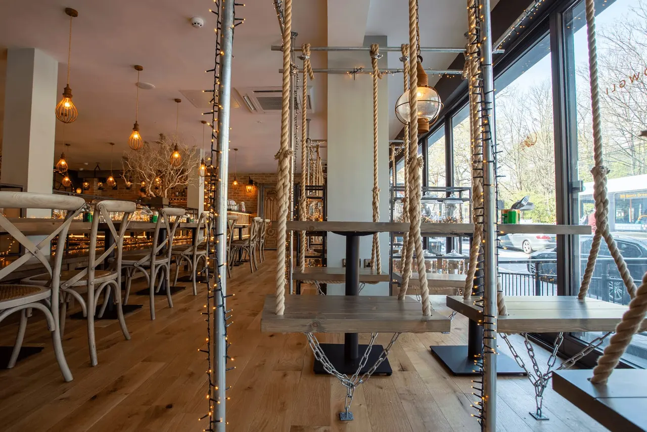 Industrial-style restaurant interior with rope-suspended tables, wooden chairs, brass pendant lights and large windows overlooking the street.
