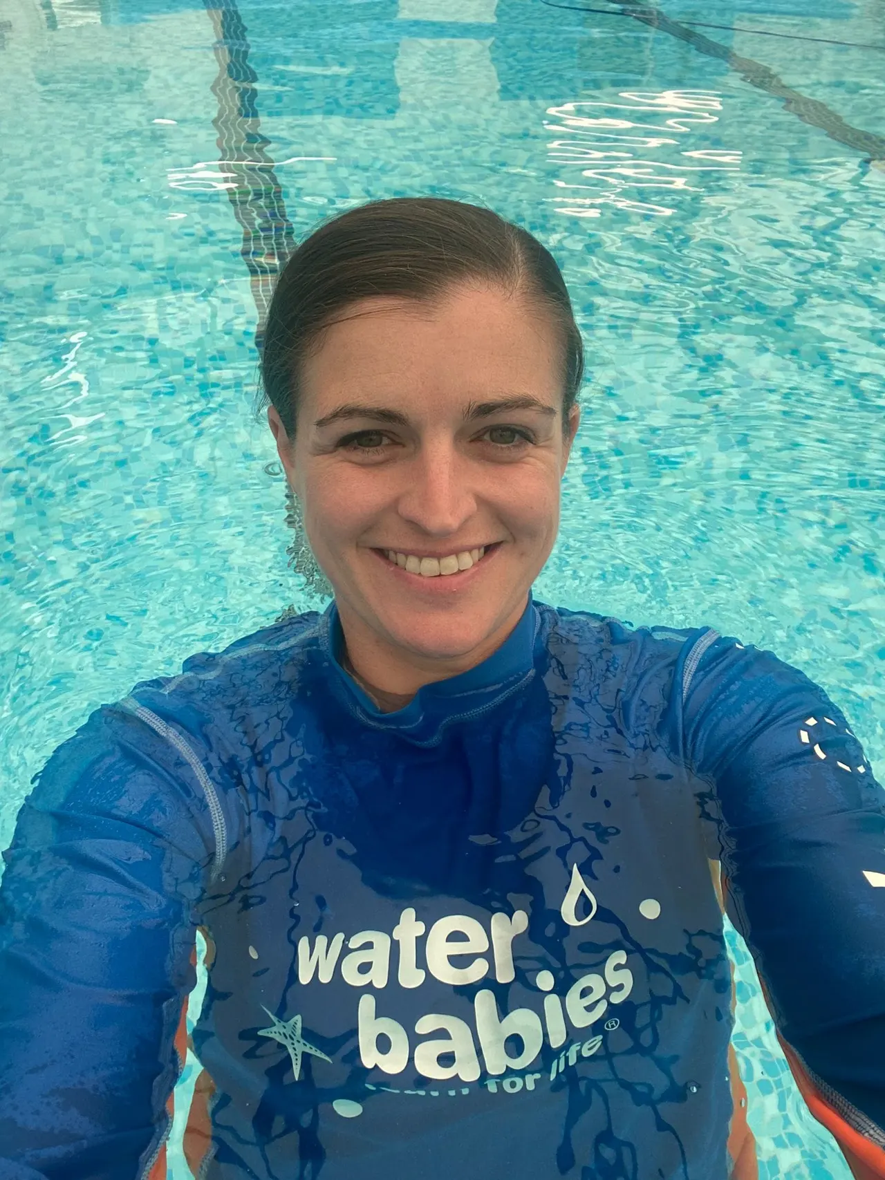 Woman in blue rash vest taking selfie at edge of indoor swimming pool.