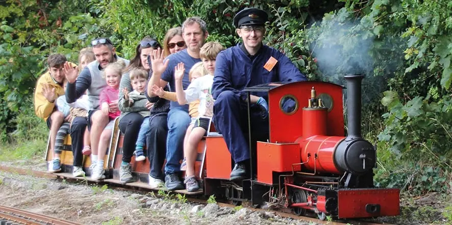 Adults and children ride on a small red and black miniature steam train surrounded by greenery.