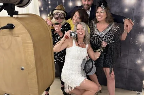 A group of five people posing together in a photo booth with a sparkly backdrop, holding props and drinks.