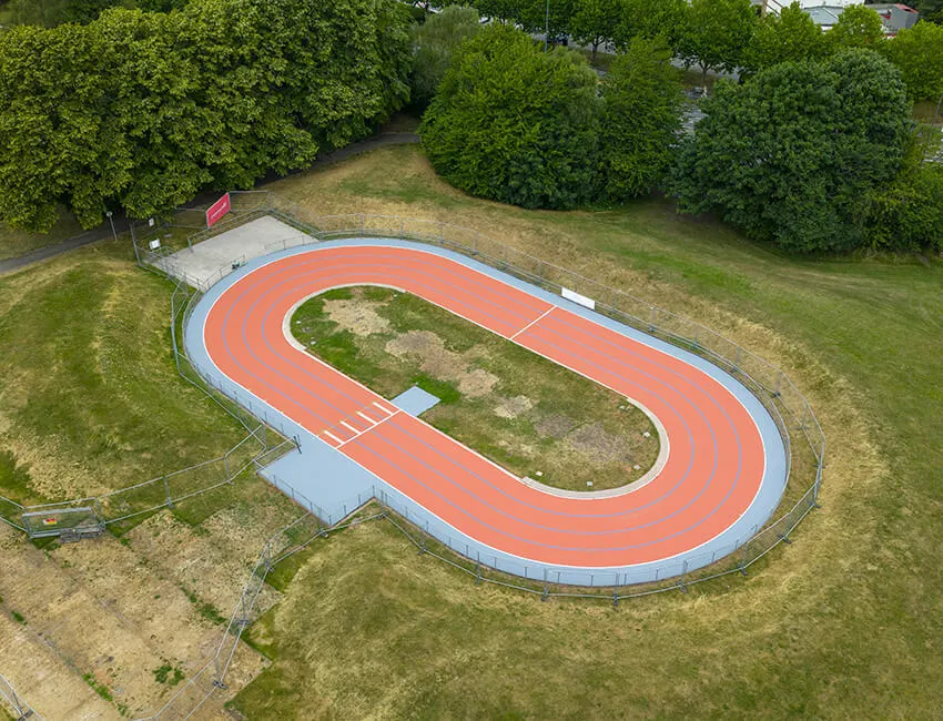 Aerial view of an outdoor athletics track with red running surface and blue lanes, surrounded by grass and trees.