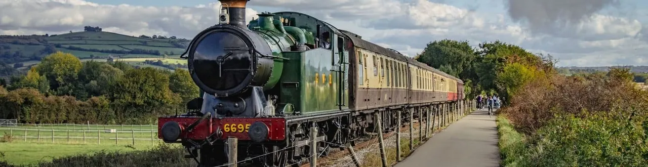 Heritage steam locomotive with cream and green passenger carriages at a rural railway station, with visitors boarding.