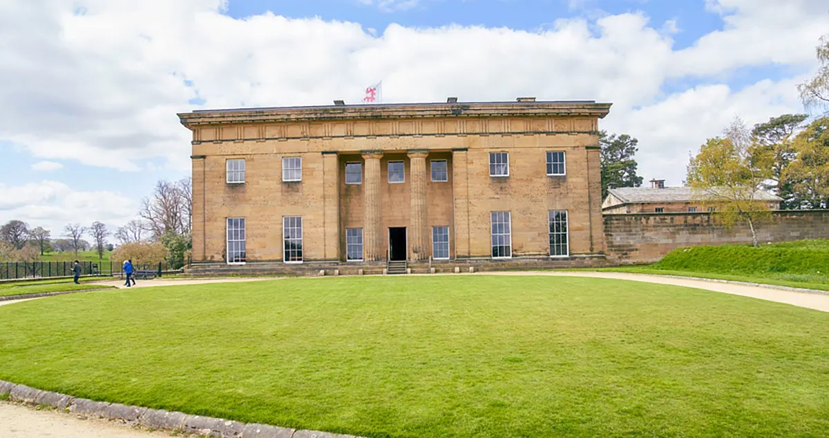 Grand stone country house with columned façade set behind open lawns and pathways, with visitors strolling the grounds on a cloudy day.