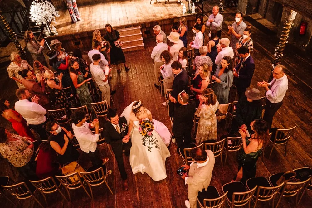 Bride and groom dancing with wedding guests in a circle on a wooden floor in a rustic hall.