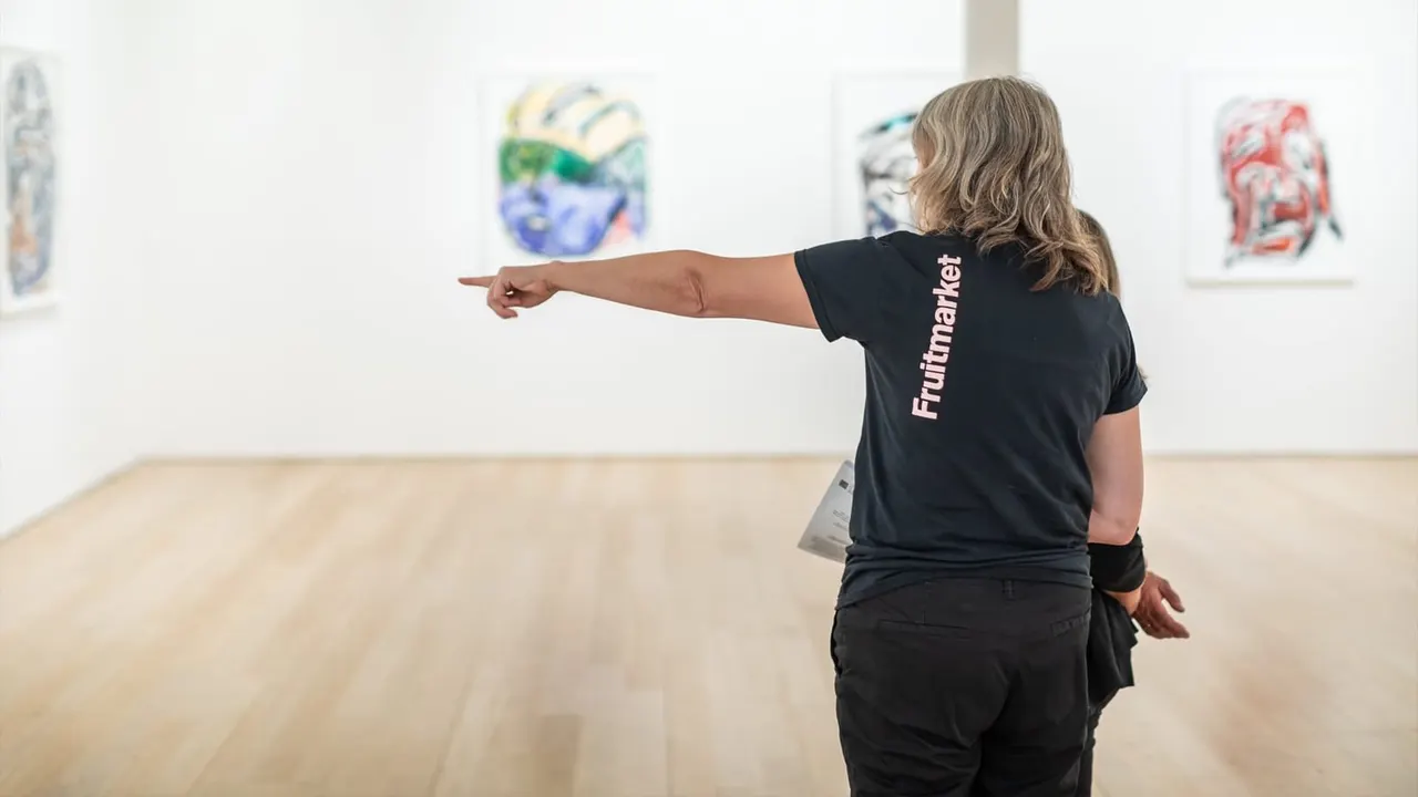 A staff member points towards colourful artworks displayed on white gallery walls in a bright, minimalist exhibition space.
