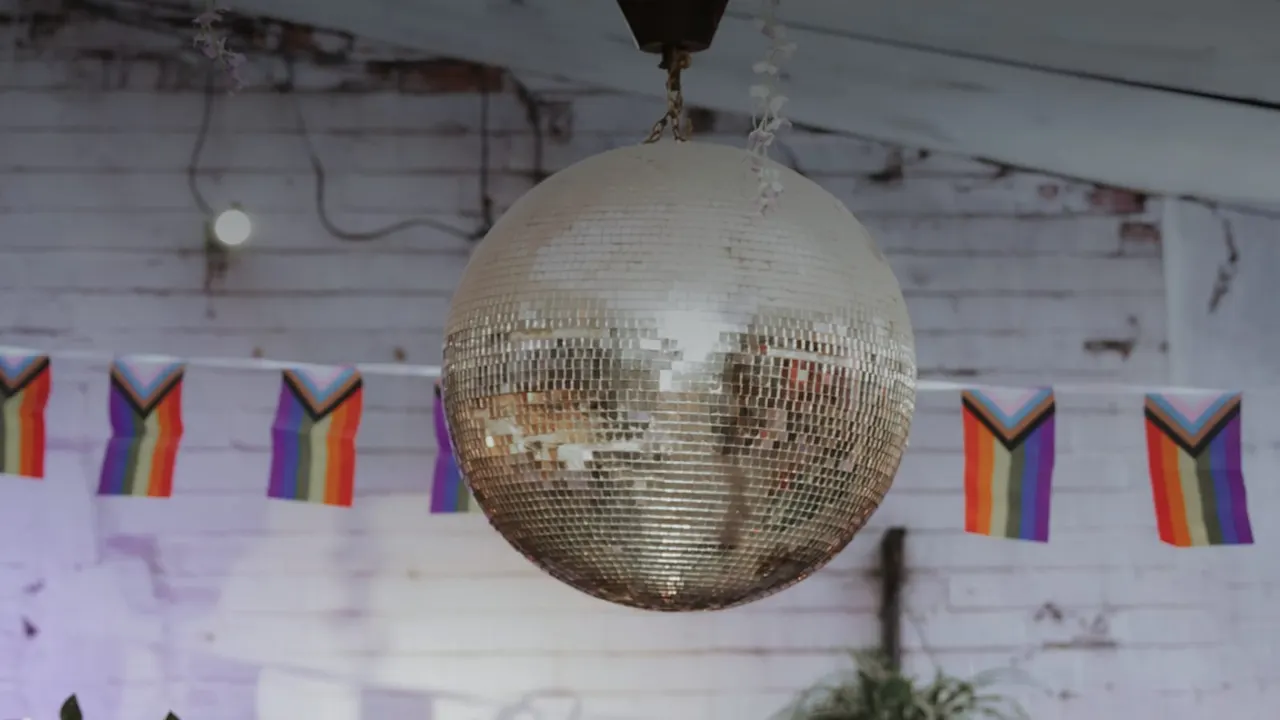 Disco ball hanging from ceiling with rainbow pride flags strung along white brick wall behind.