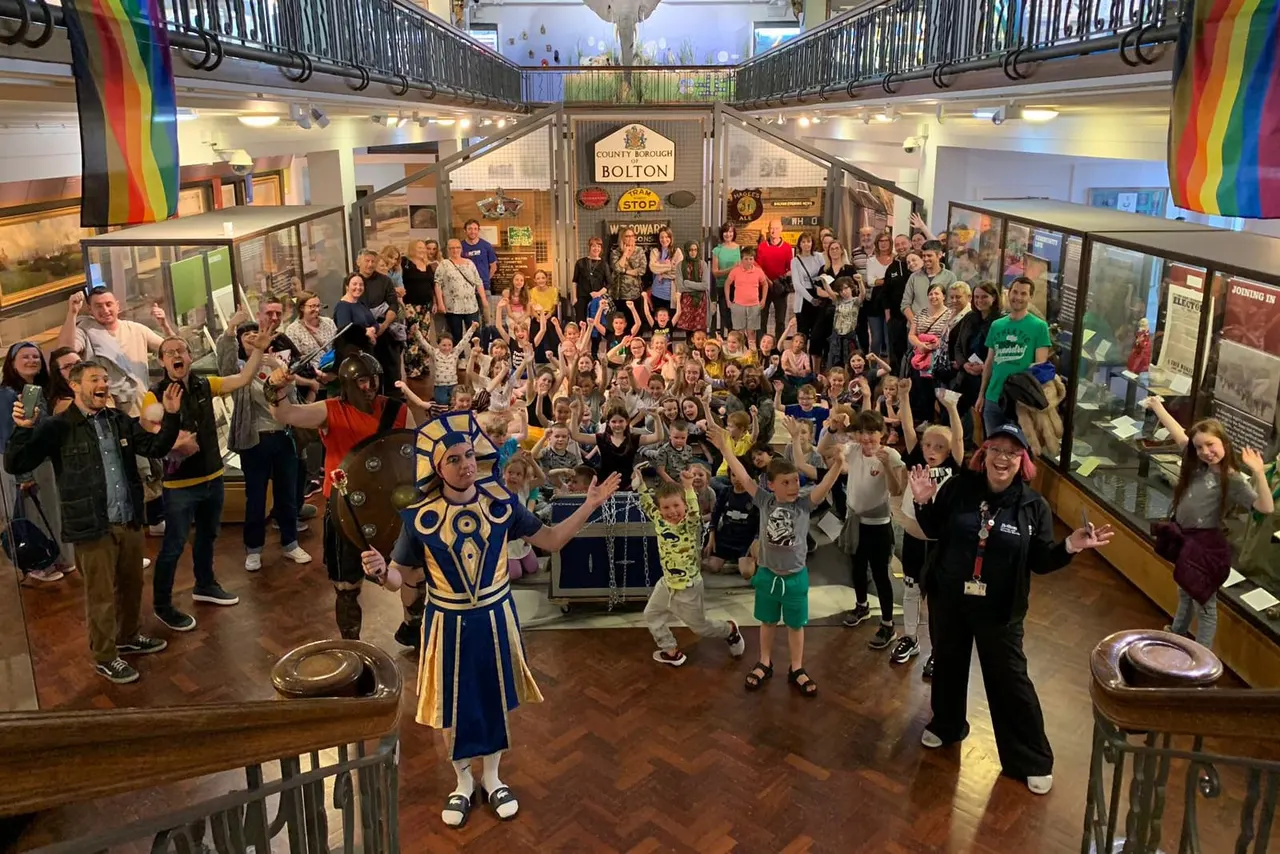 A large group of visitors of all ages gathered in a museum gallery space, with a costumed performer and staff member leading an interactive activity.