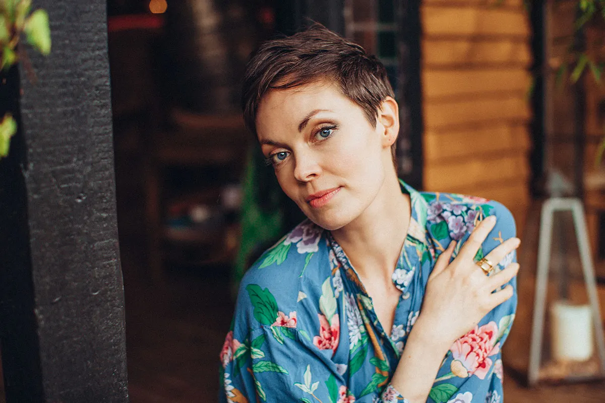 Woman with short brown hair wearing a blue floral shirt, leaning against a dark pillar indoors.