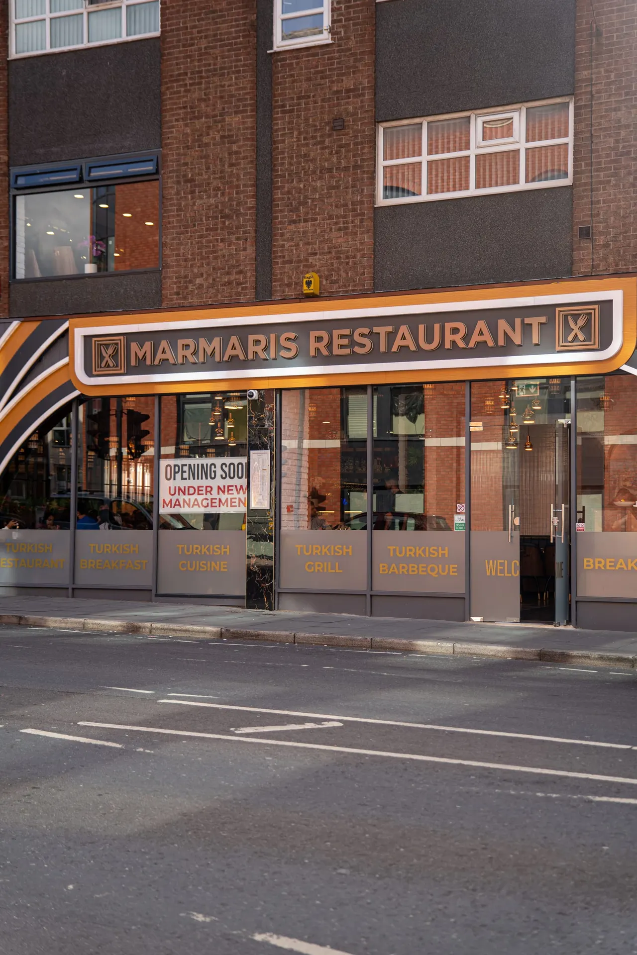 Storefront of a Turkish restaurant with orange and white signage on a brick building, showing windows and an "opening soon under new management" notice.