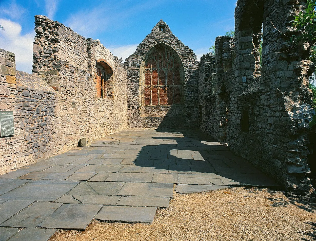 Ruins of a stone medieval church interior with Gothic arch window and ivy-covered walls, clear blue sky above.