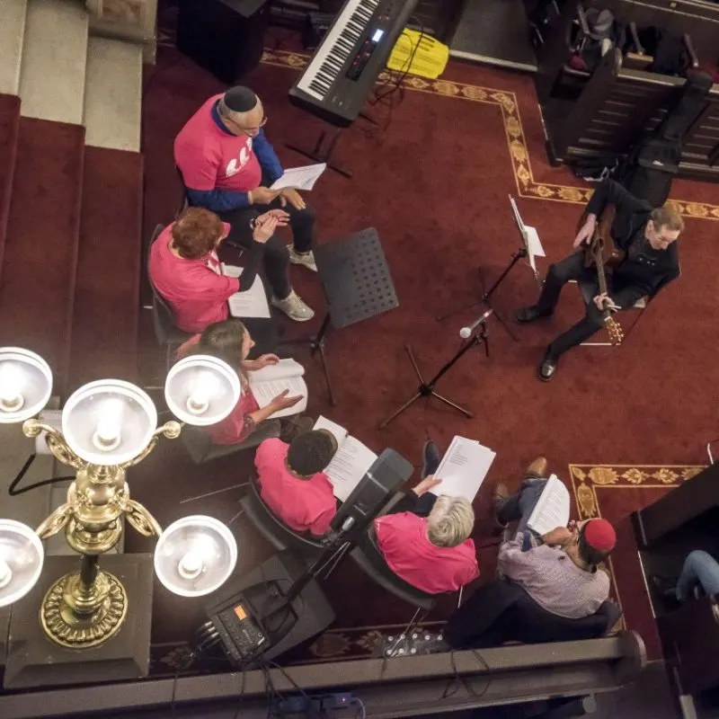 Choir members in pink shirts rehearsing with sheet music in an ornate interior space with stage lighting.