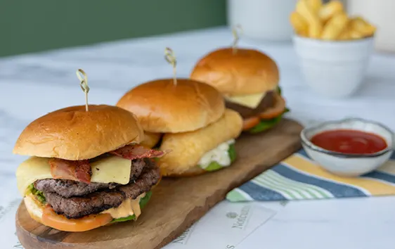 Three cheeseburgers with toppings arranged on a wooden board, served with fries and ketchup.
