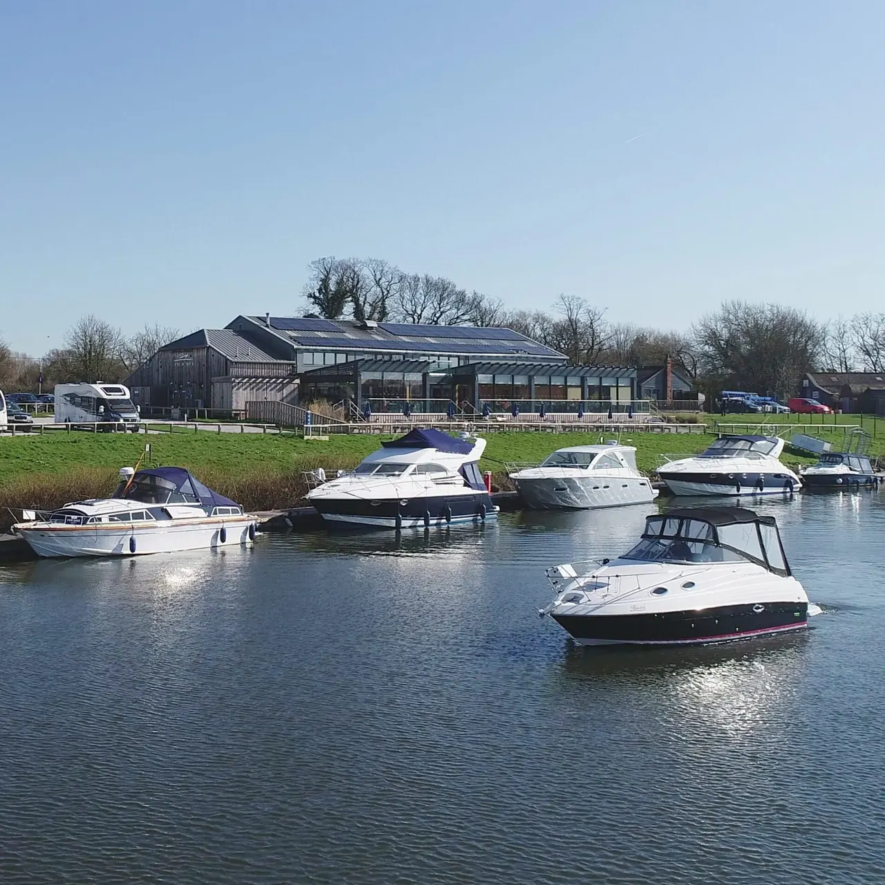 Several moored boats on water with a modern waterside leisure building and green fields behind.