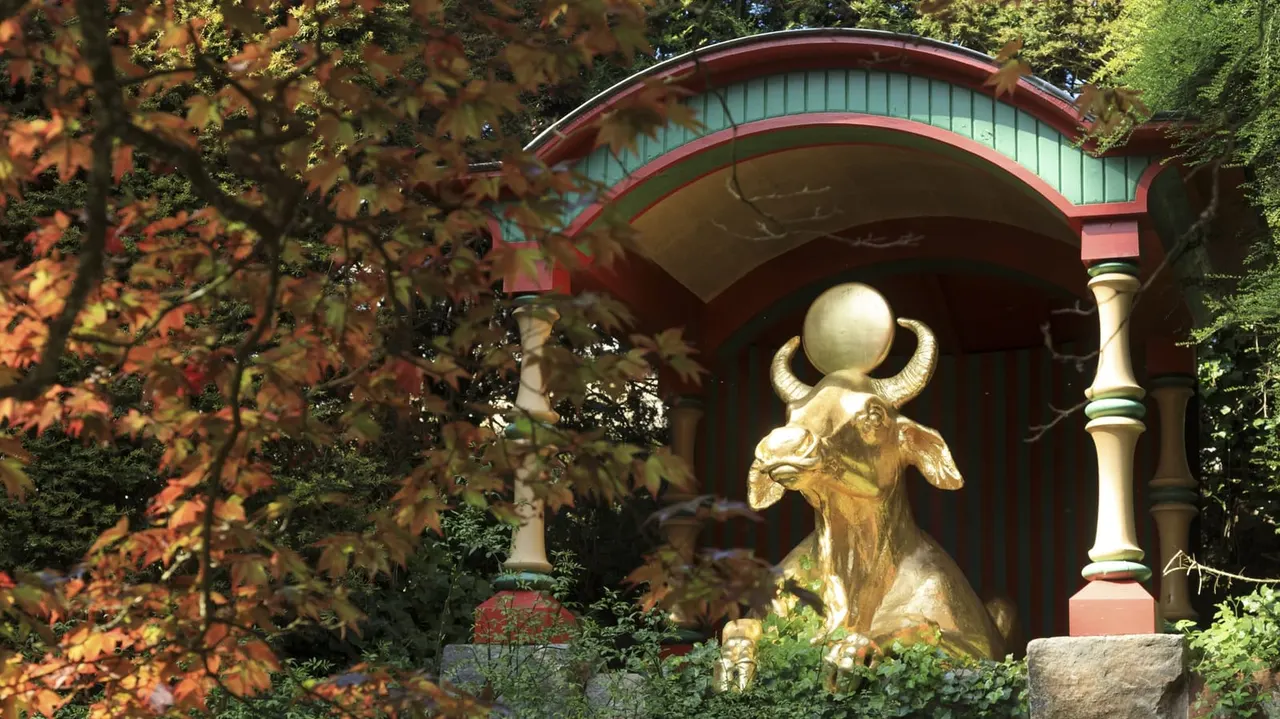 Gilded bull statue beneath a curved red and green pavilion, framed by autumn foliage and climbing ivy.
