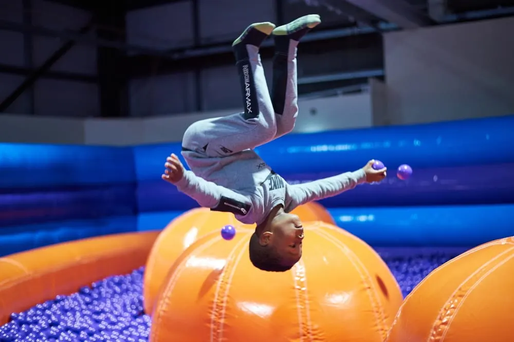Person doing a backflip over large orange inflatables in an indoor trampoline park with blue padding.