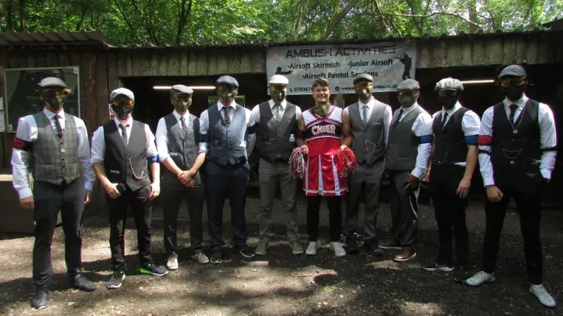 A line of staff members wearing waistcoats and flat caps stand in front of a wooden shelter, one holding a red airsoft jersey.