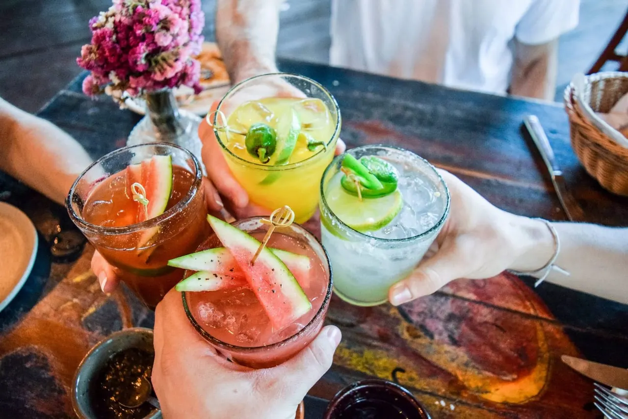 Three people holding colourful cocktails garnished with watermelon and cucumber slices at a table with pink flowers.