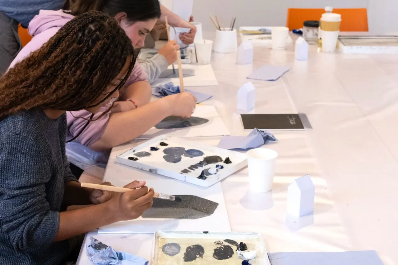 Children and adults painting with brushes at a long table during an art workshop session.