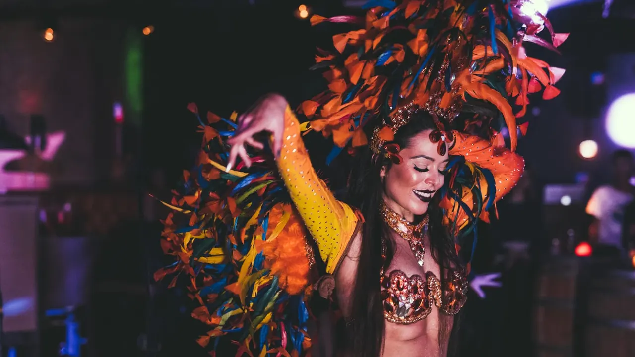 Performer in sparkly carnival costume with elaborate orange and yellow feathered headdress dancing in a nightclub with coloured lights.