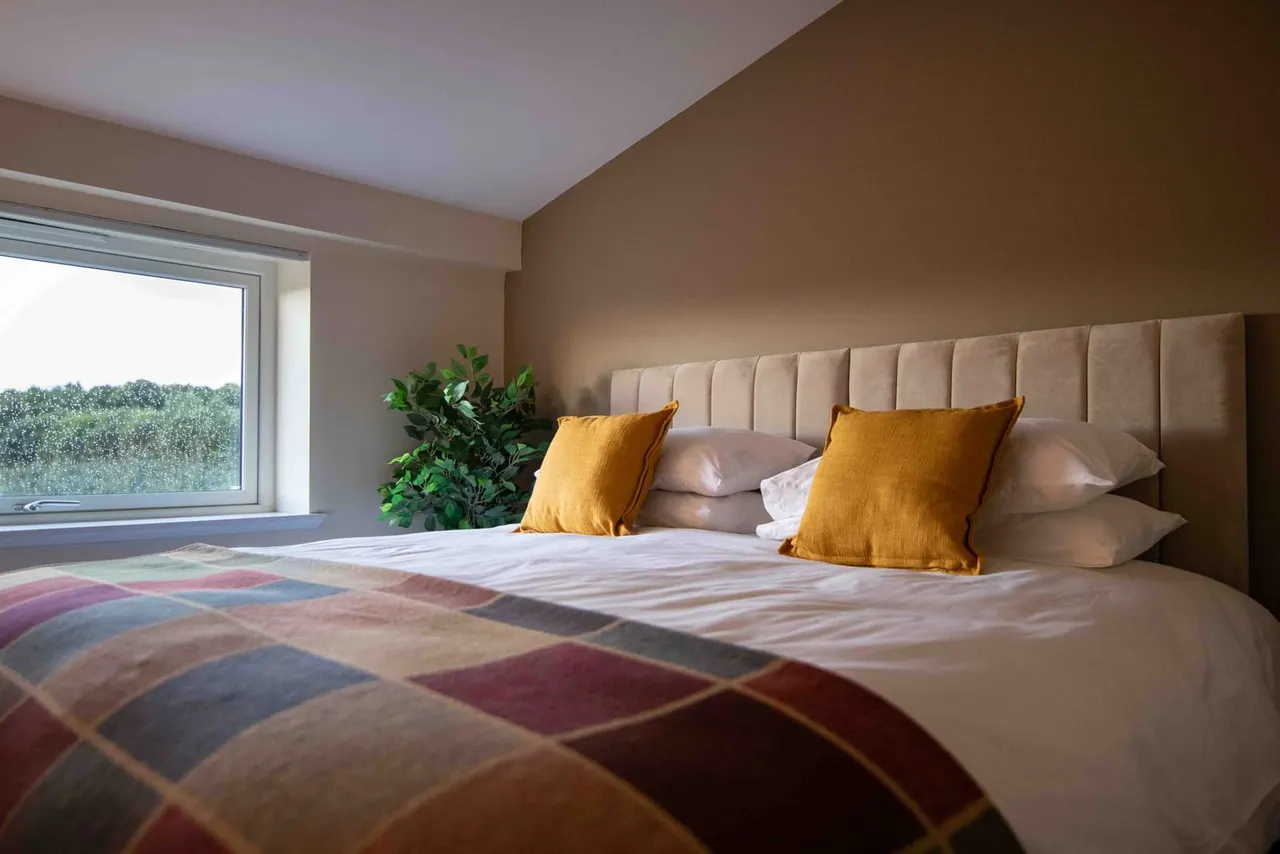 Modern bedroom with double bed, beige upholstered headboard, yellow cushions, and countryside views through a large window.