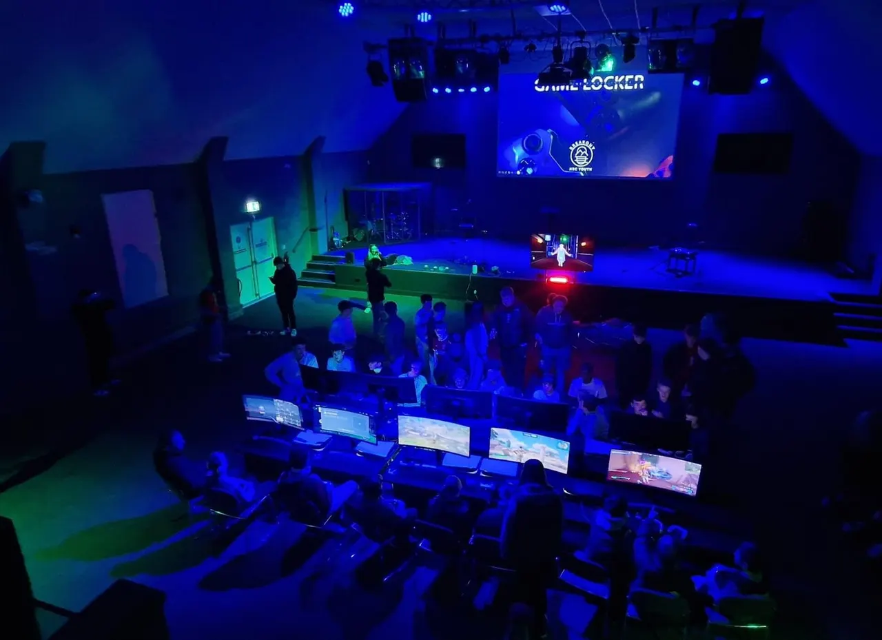Indoor gaming arena with groups of people at computer stations and a stage area lit in blue and green neon lighting.