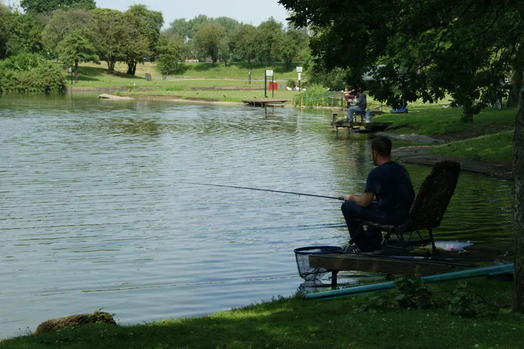 Anglers fishing from the riverbank with trees and green parkland surrounding the water.