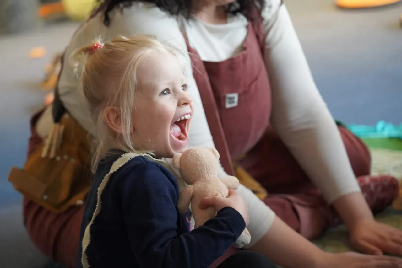 Young child holding a teddy bear, laughing with an adult carer during an indoor activity session.