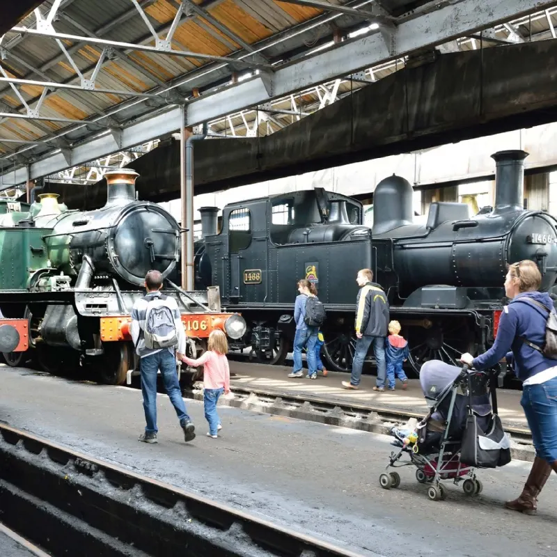 Visitors and families exploring historic steam locomotives under a railway depot shed.
