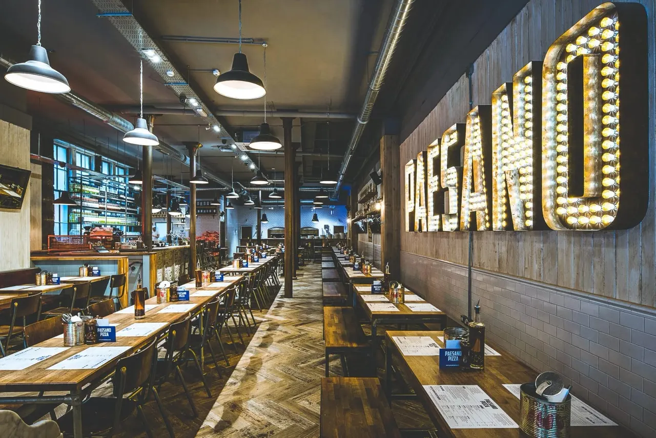 Modern pizzeria dining room with wooden tables, black chairs, industrial pendant lights, and illuminated signage on the wall.
