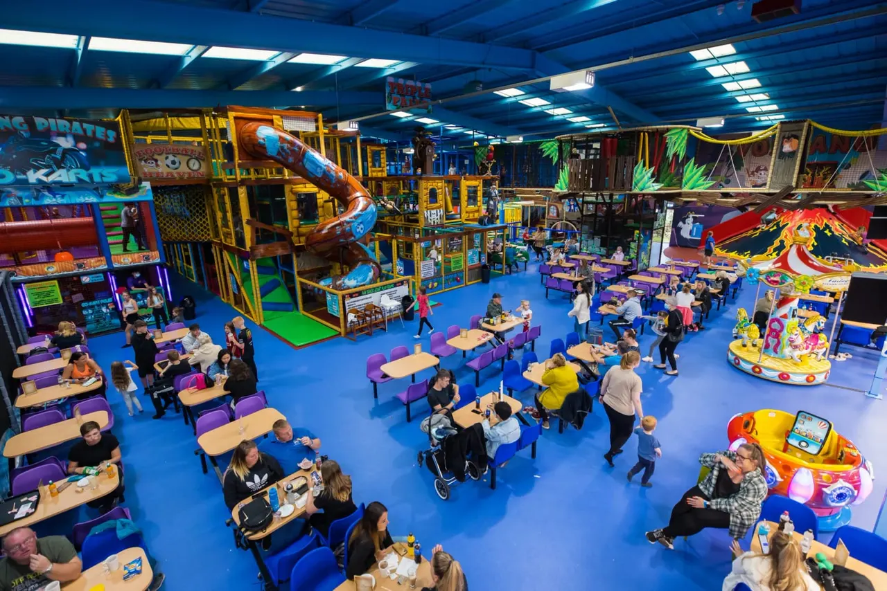 Indoor children's play venue with families seated at tables, kids using soft play structures, slides and rides under bright blue lighting.