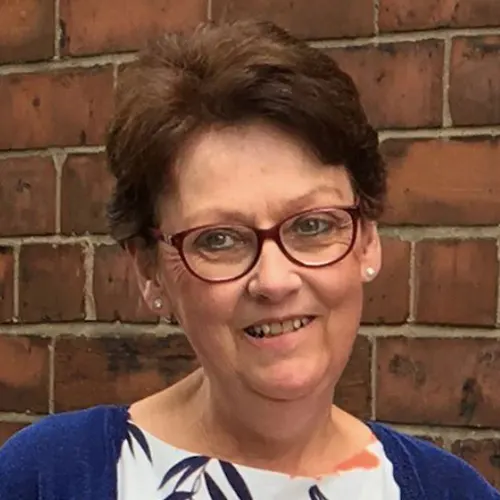 This is a PHOTOGRAPH, not a designed graphic. Here is the alt text:

Woman wearing glasses and blue top smiling at camera against brick wall background.