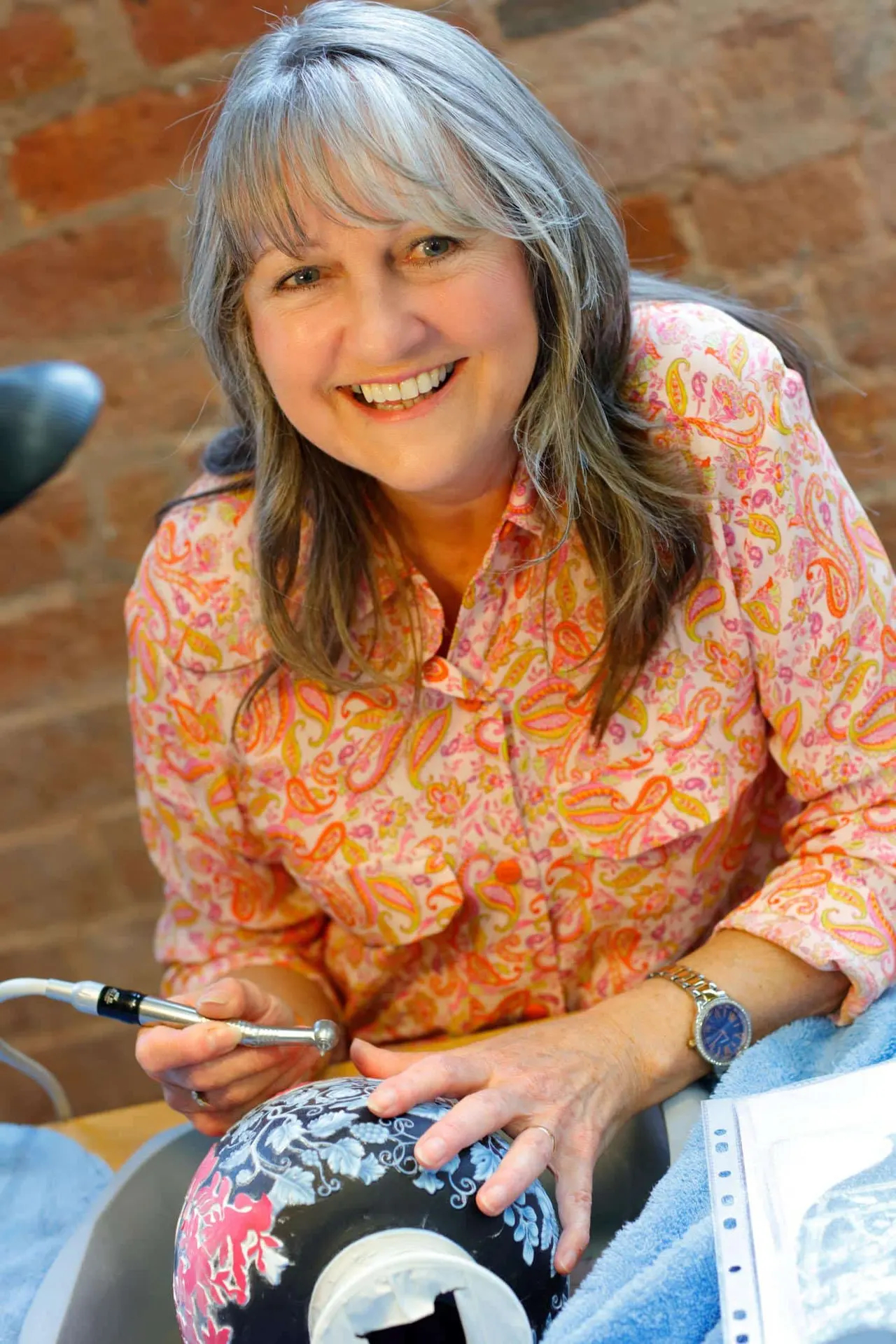 Woman with grey hair smiling at camera whilst holding a paintbrush and decorating a piece of glassware.